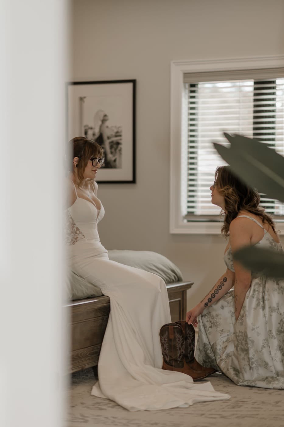A bride sits on the edge of the bed in her gown talking with a bridesmaid kneeling beside her, cowboy boots on the floor between them