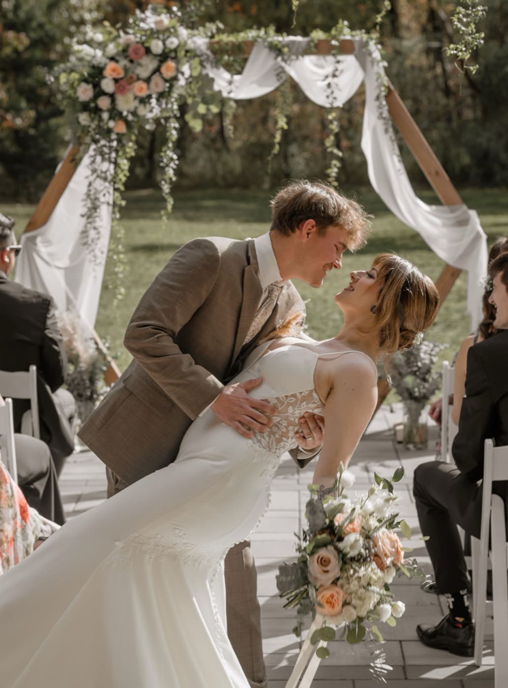 A groom dips his bride for a kiss at the end of an outdoor ceremony, framed by a floral arbour