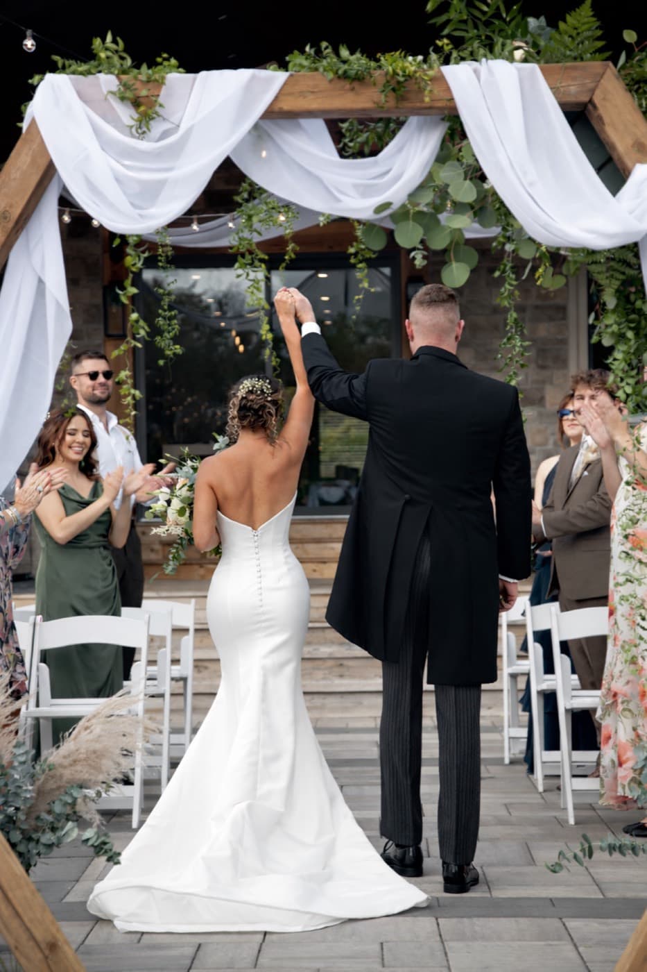 A newly married couple walk back down the aisle hand in hand beneath a draped wooden arbour, guests applauding on either side