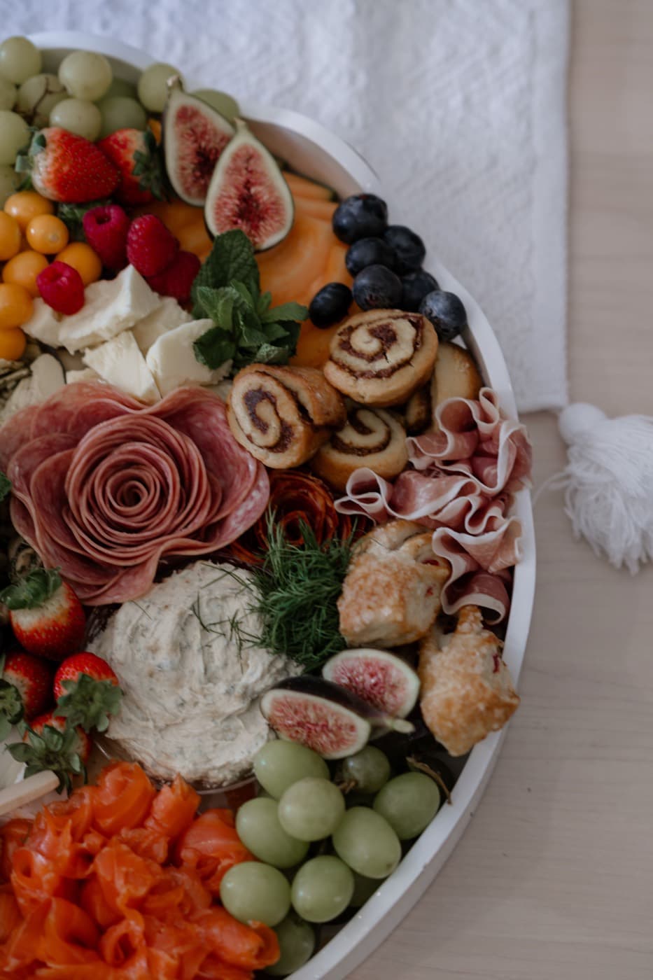 A charcuterie board seen from above with a salami rose at its centre, figs, smoked salmon, grapes, soft cheese and pastry bites