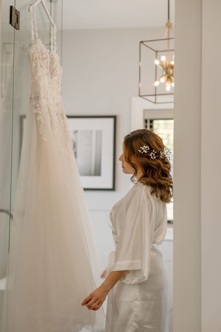 A bride in a robe stands beside her beaded tulle gown where it hangs, reaching out to touch the skirt