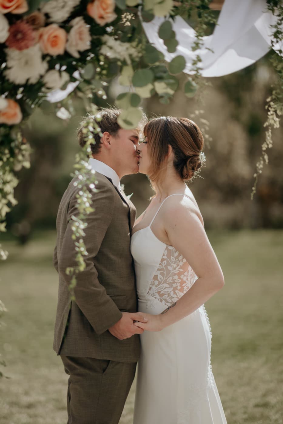 A couple kiss under the floral arbour at the end of their ceremony, the groom in a brown suit and the bride in a lace gown