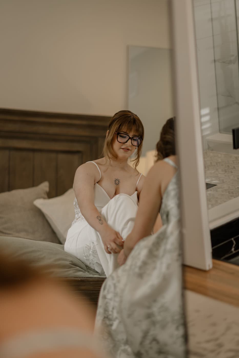 A bride sits on the bed in her gown while a bridesmaid helps with the hem, morning light across the suite