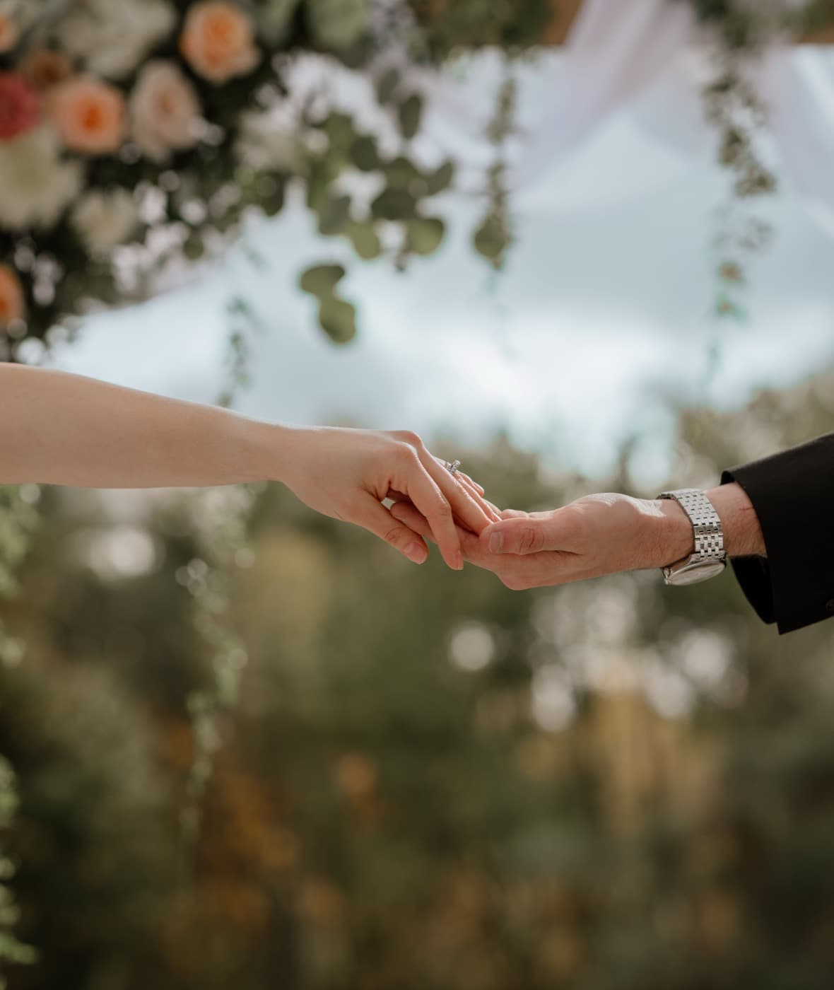 Two hands reaching for one another during the ceremony, the flowers of the arbour blurred above them