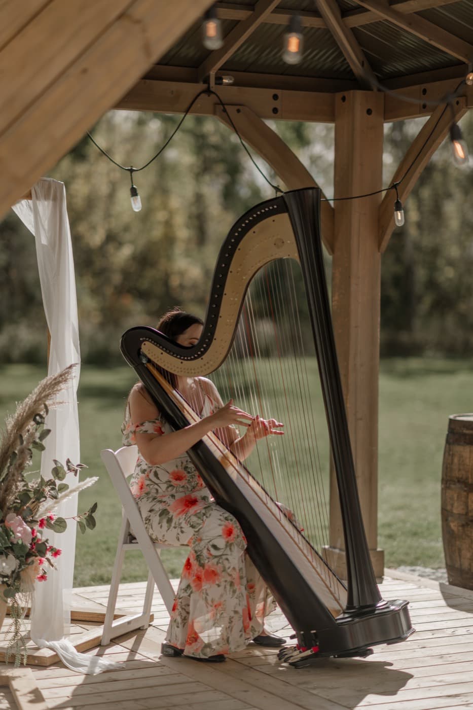 A harpist in a floral dress plays beneath the wooden gazebo, string lights overhead and the lawn beyond