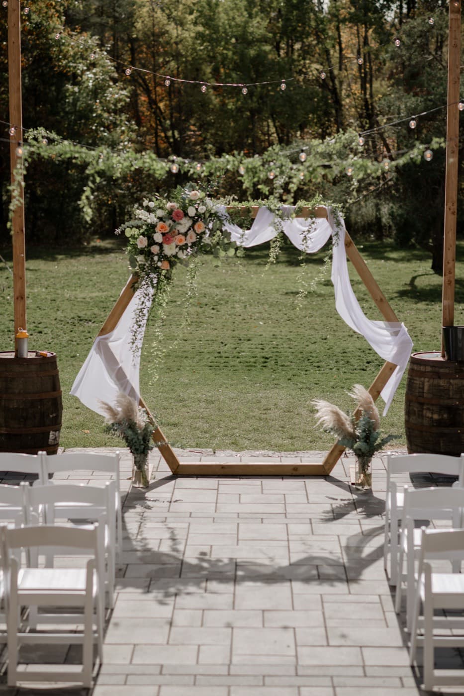 A hexagonal wooden ceremony arbour on the patio, dressed with peach and white roses and white drapery, rows of white chairs facing it and string lights strung above the lawn