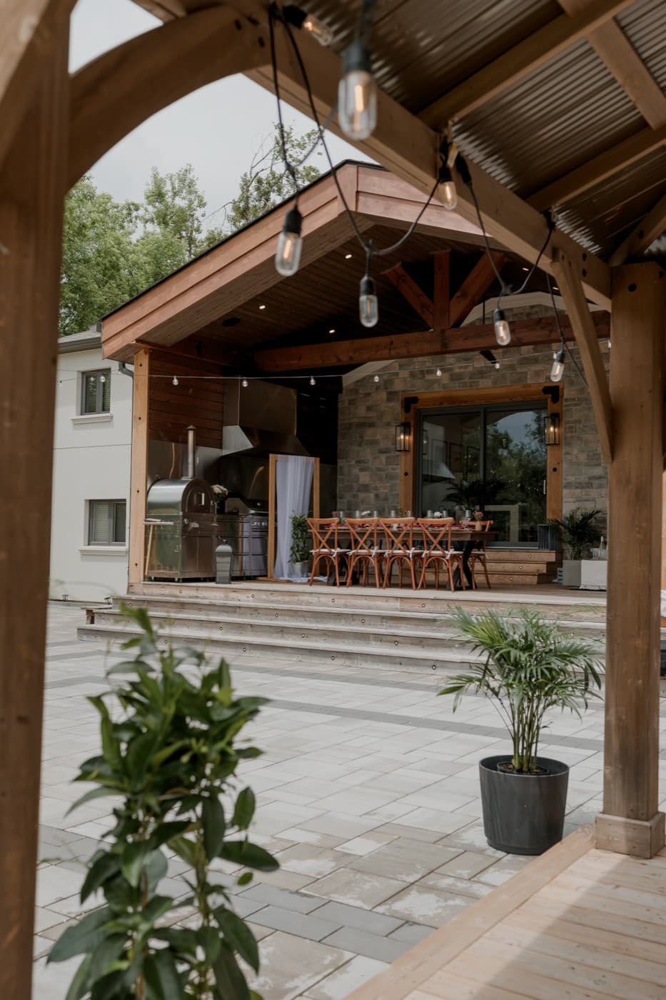 The covered pavilion and its long set table seen through the timber pergola, string lights hanging across the stone patio