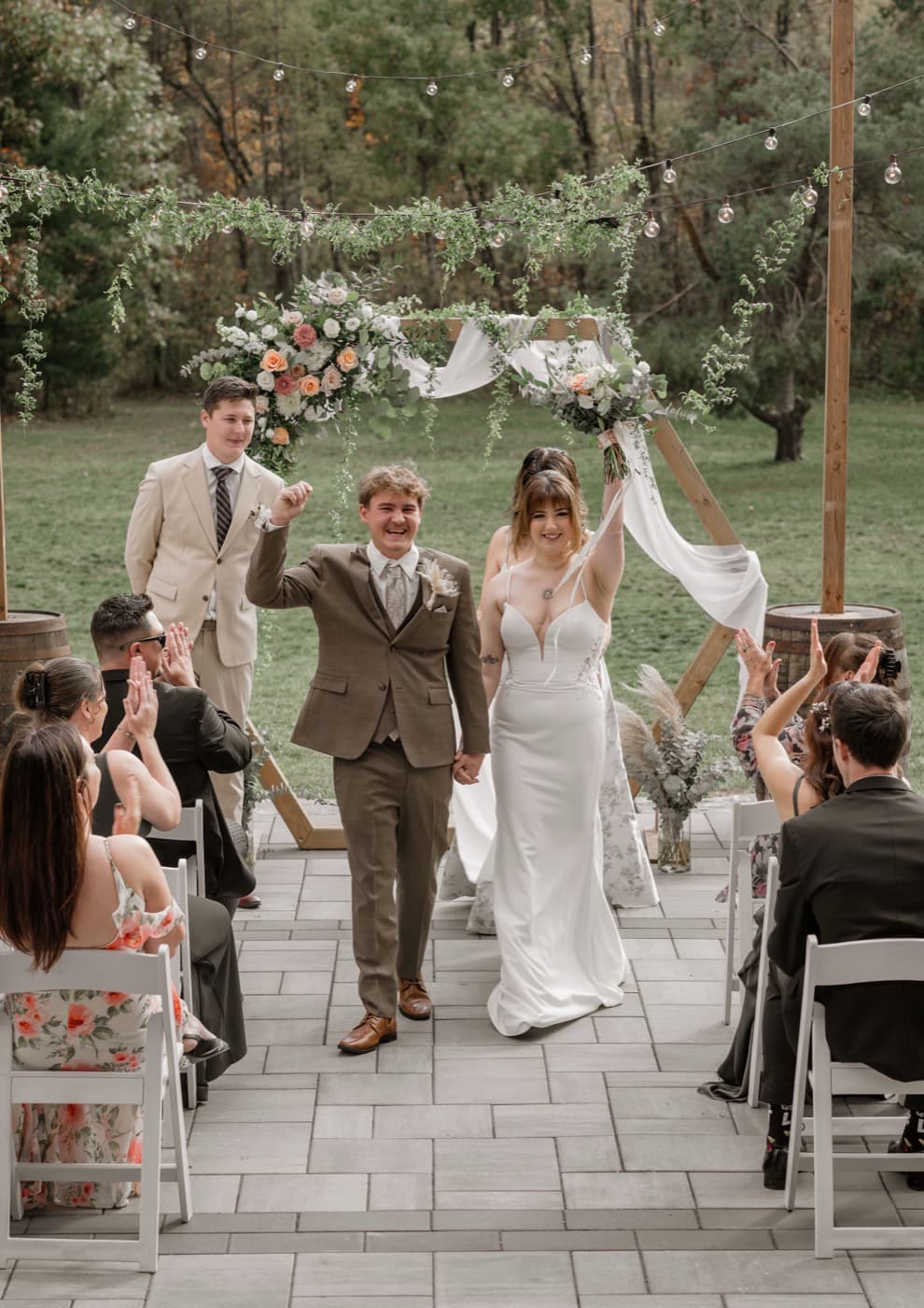 A couple walk back up the aisle with their arms raised, guests applauding from white chairs on both sides