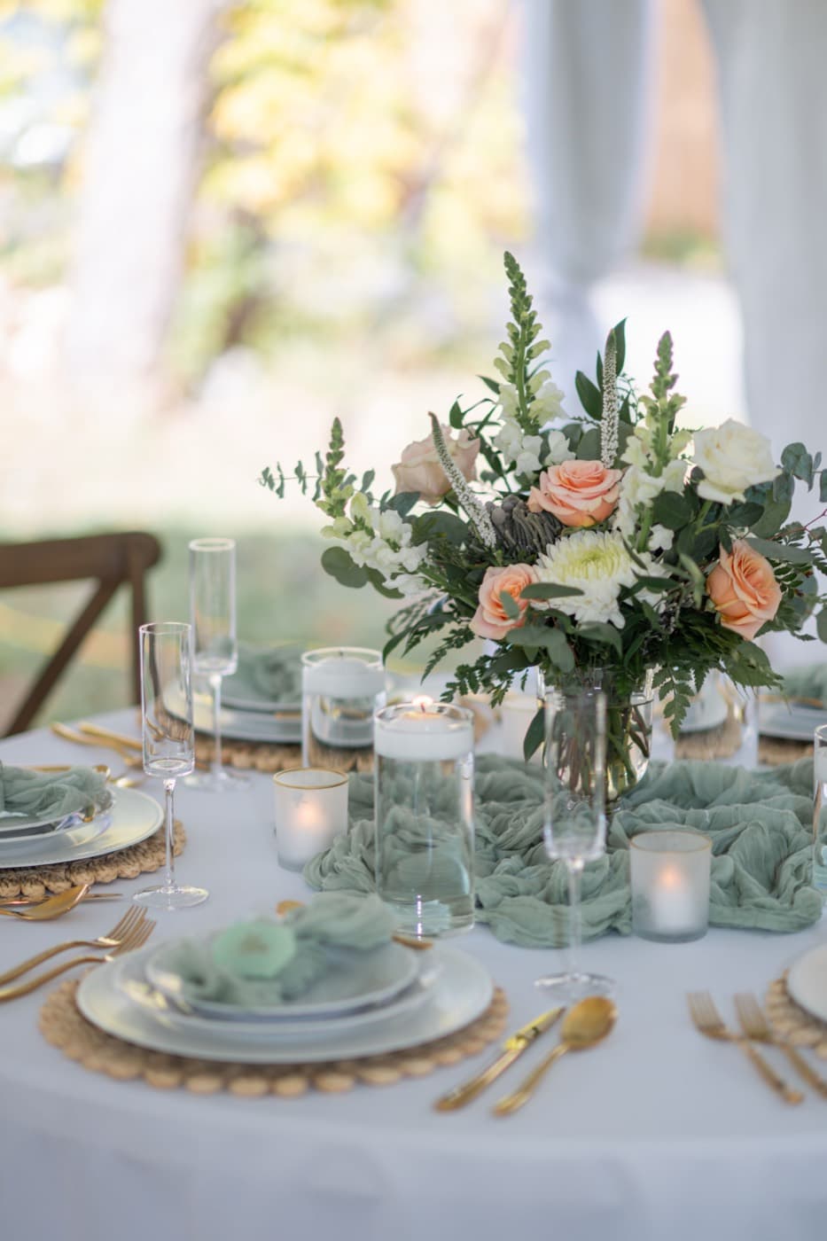 A round table with a sage gauze runner, rattan chargers, gold cutlery, floating candles and a peach and white floral centrepiece