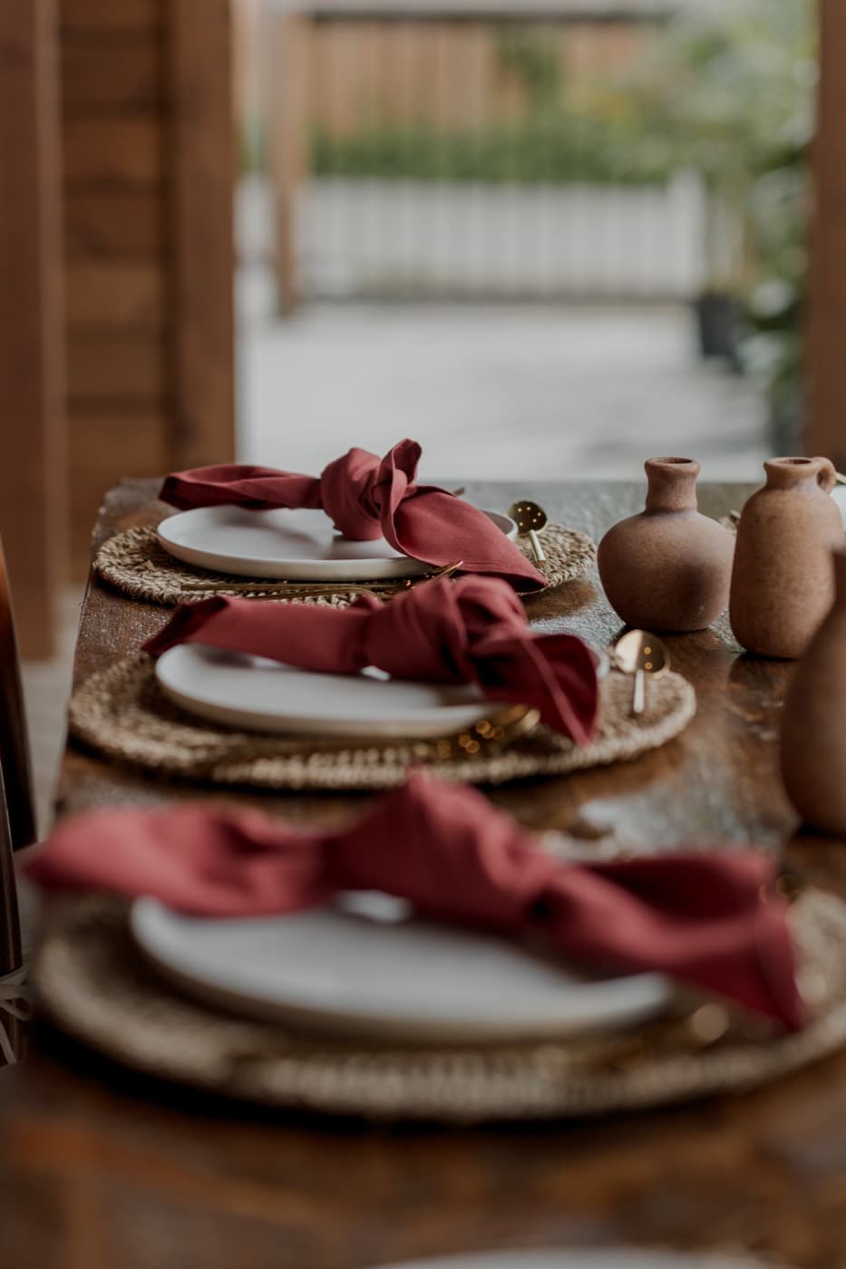 Three place settings in a row, rust napkins knotted on white plates over woven chargers beside terracotta bud vases