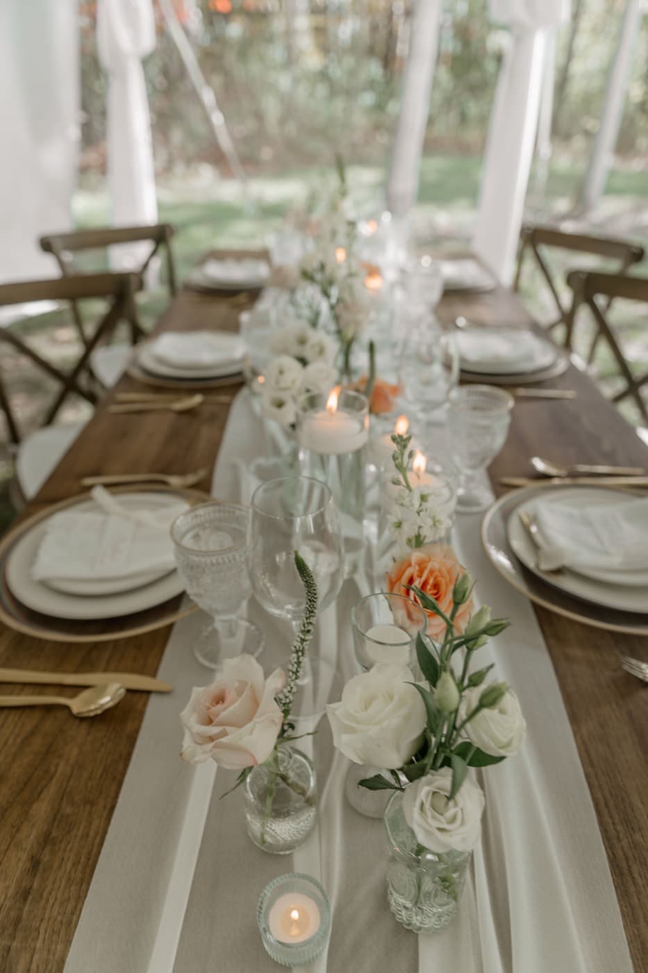 Looking down a reception table over a gauze runner, bud vases of peach and white roses, floating candles and gold cutlery