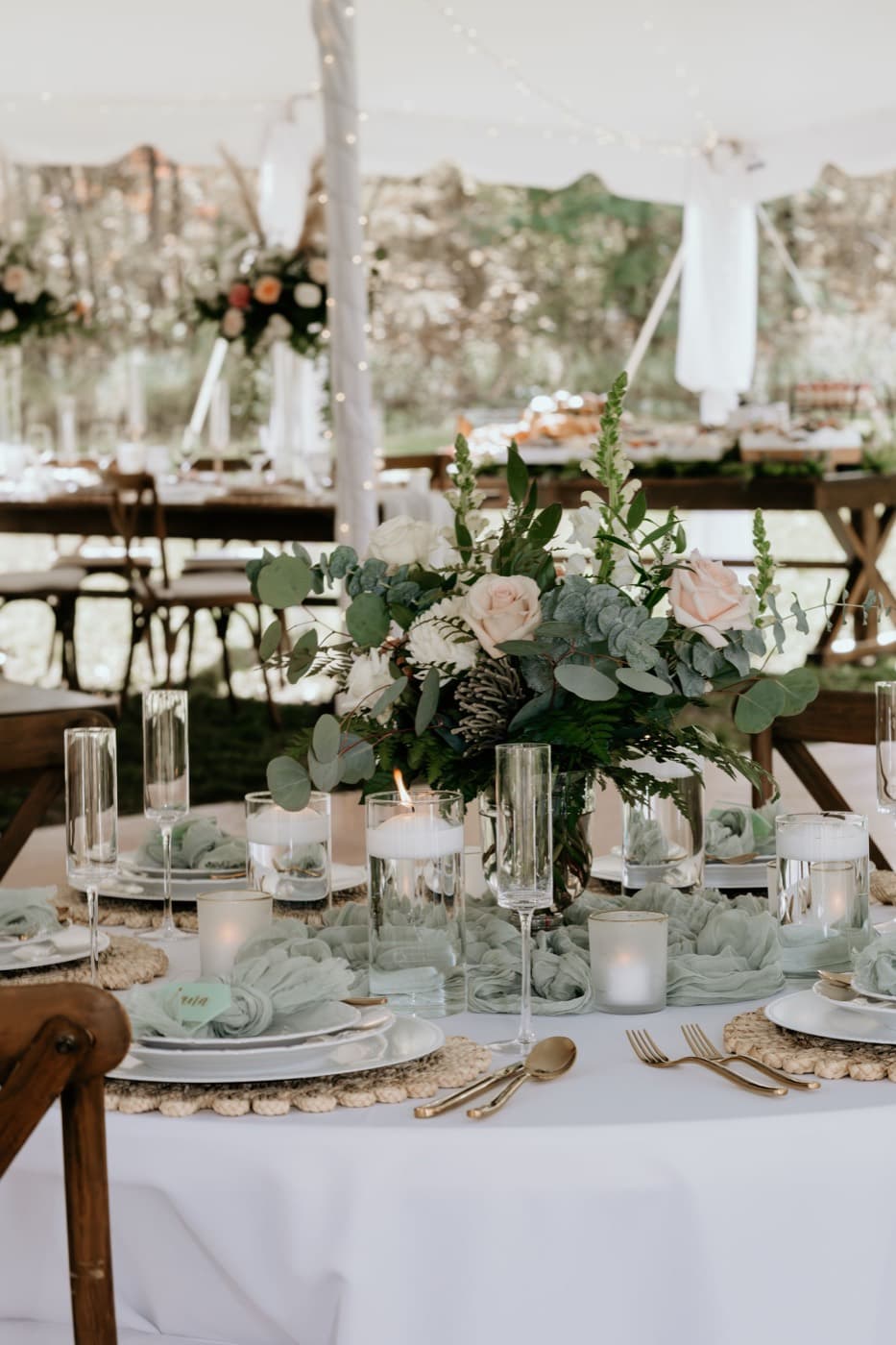 Round tables laid across the tent with sage runners, floral centrepieces and string lights, autumn woods beyond the open sides