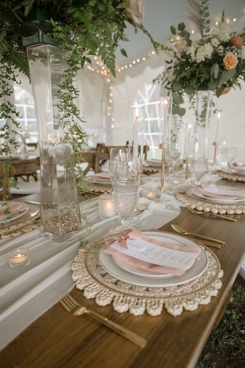 A place setting under the tent with gold cutlery, a macrame charger, a blush napkin and a menu card, tall greenery centrepieces behind