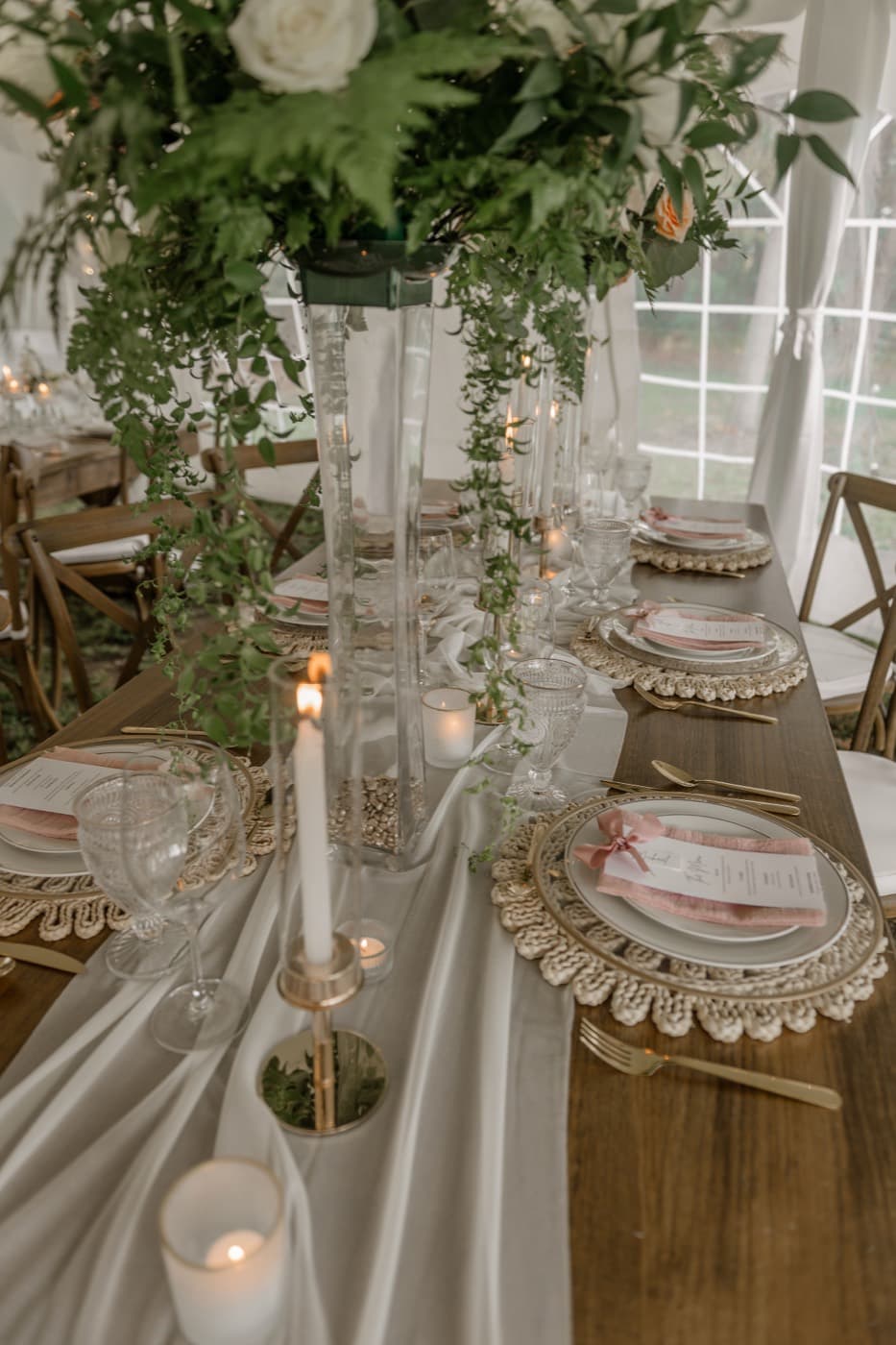 A long tented table at dusk with tall glass vases of ferns and trailing greenery, taper candles and macrame chargers