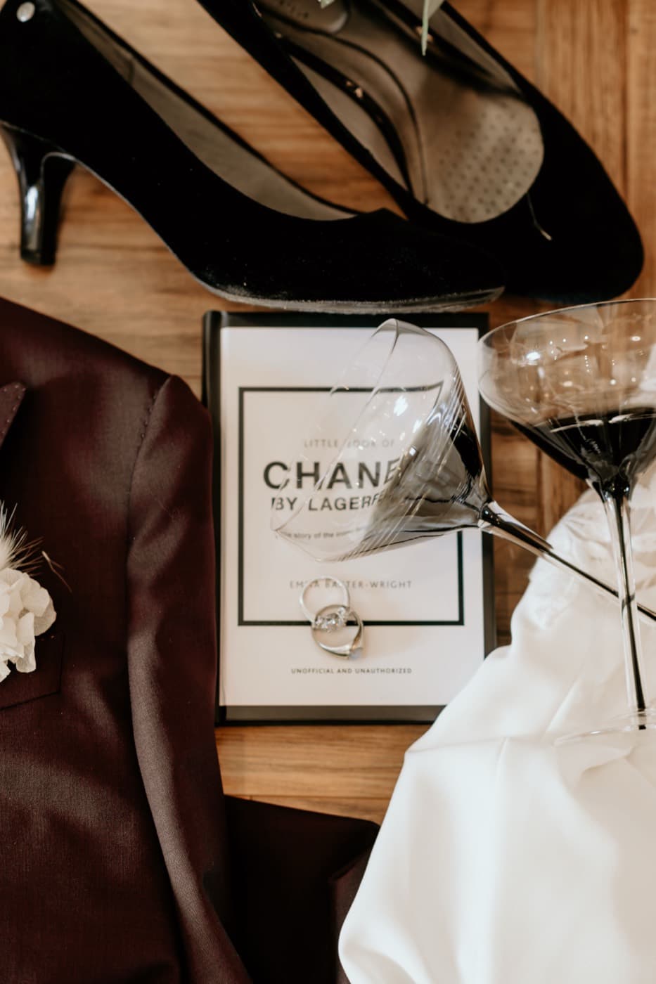 Wedding day details laid out on a wooden floor: black velvet heels, a burgundy jacket with its boutonniere, coupe glasses and the rings