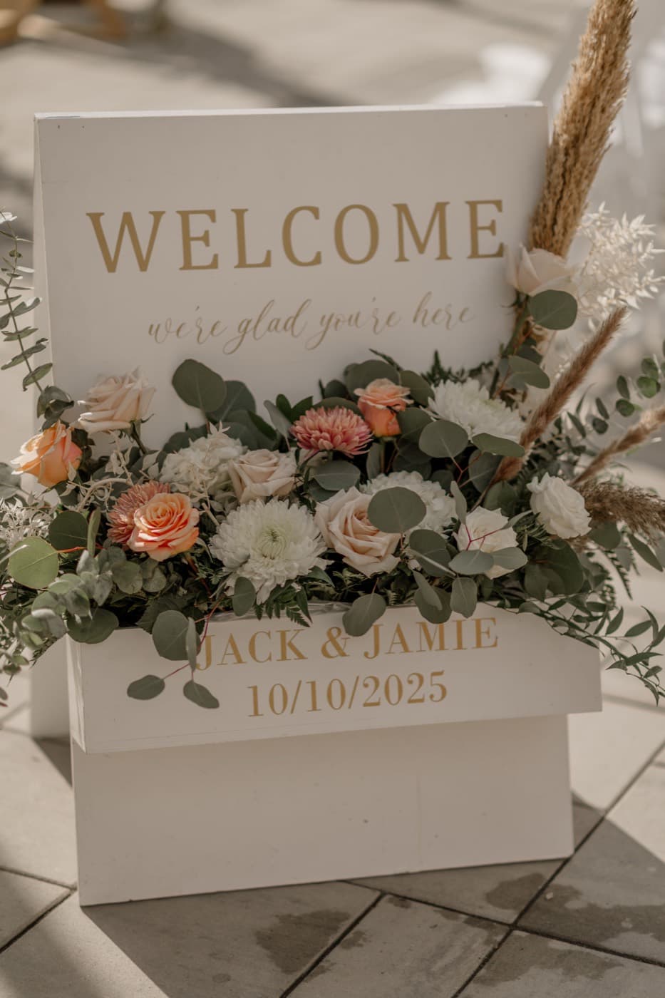 A white welcome sign on the patio reading "we’re glad you’re here", banked with peach roses, white blooms, eucalyptus and pampas