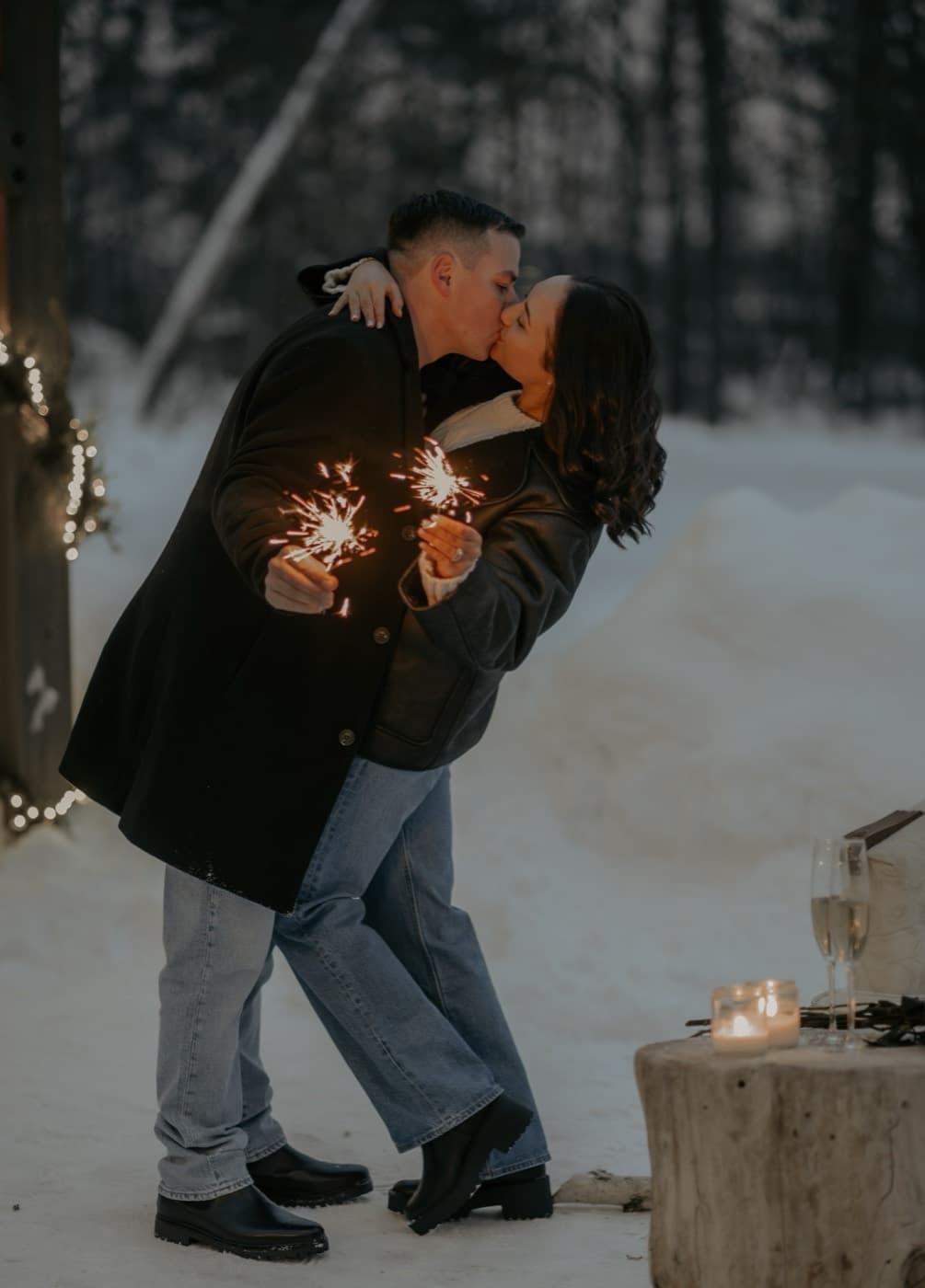 A couple kiss in the snow at dusk holding sparklers, with champagne and a candle on a tree stump beside them